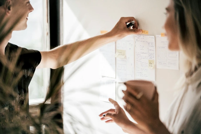 Two people collaborating on wireframe sketches pinned to a wall in a workspace
